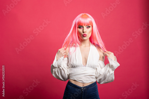 Stylish female adult in fashionable blouse and pink wig pouting confidently while holding hair. Glamorous caucasian woman standing against isolated backdrop with flirty expression.