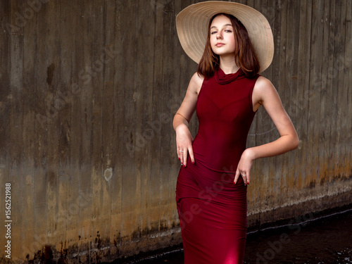 Young girl in luxurious red dress and large hat in a vogue style look posing standing in water under a bridge. Woman in a fancy outfit with slim body type. Elegant model with calm self assured look