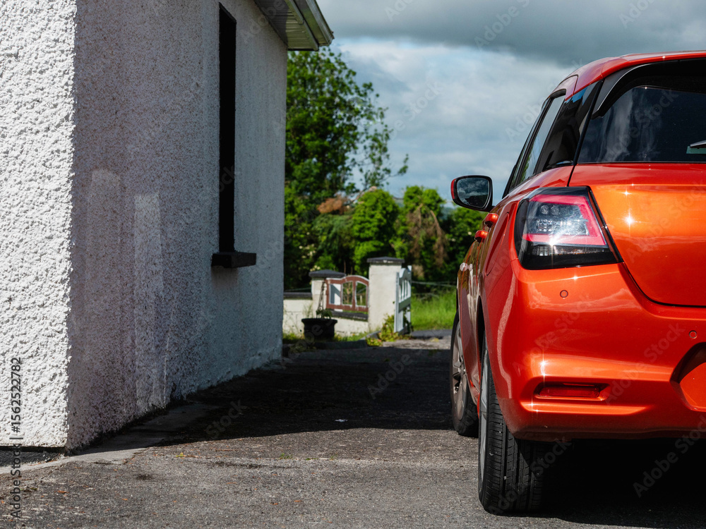 Fototapeta premium Small sport red car parked by a house with white walls, rural area in the background. Warm sunny day. Living in a country side concept. Commute and transportation theme.