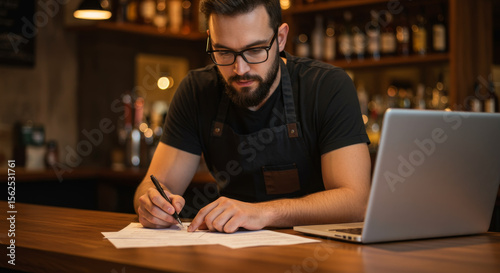 Focused bartender wearing an apron writing on paper next to a laptop with a bar in the background.