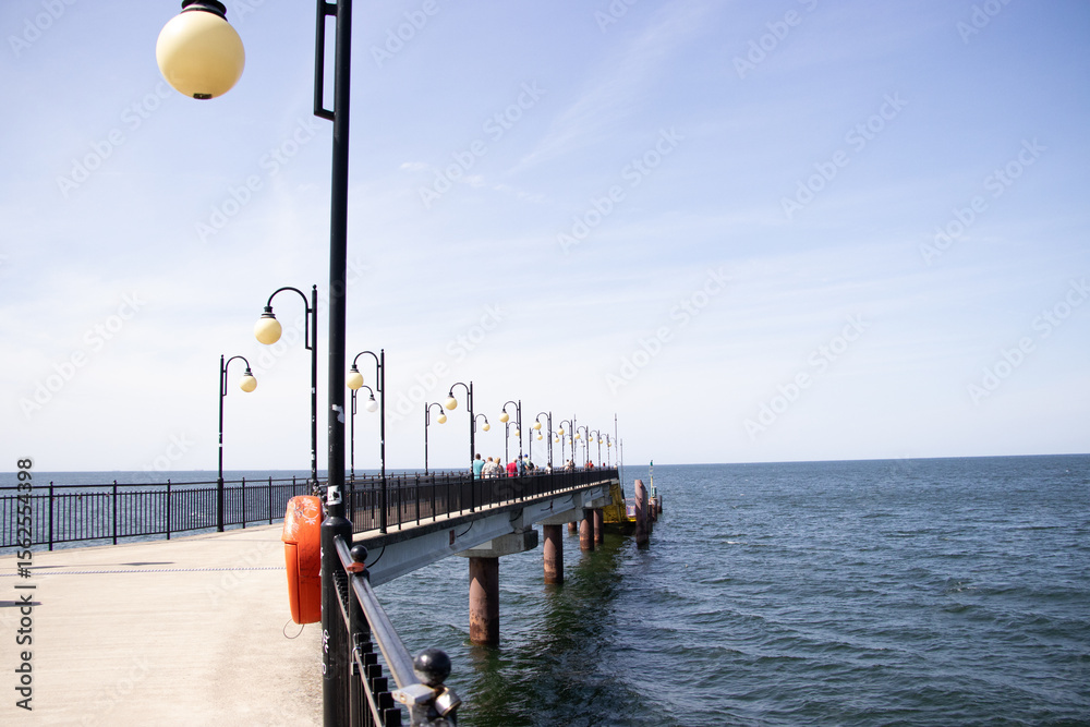 Obraz premium Metal pier with red railings and a lamppost leading to the Baltic Sea on a sunny day. Calm blue water and pale sky in the background. Photo taken in Międzyzdroje, Poland.