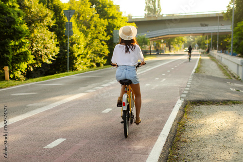 Back view of young woman riding on rent electric bike in city park at summer sunny day, full length, free space