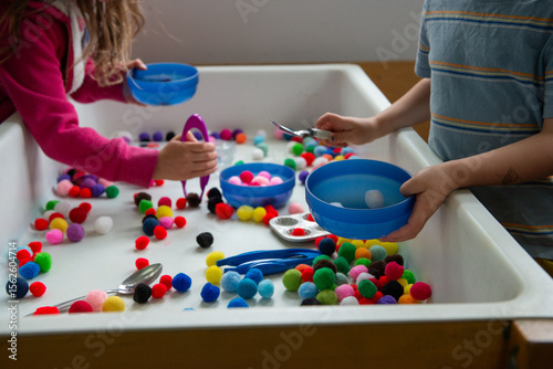 Two preschool kids play with puff balls at a sensory table