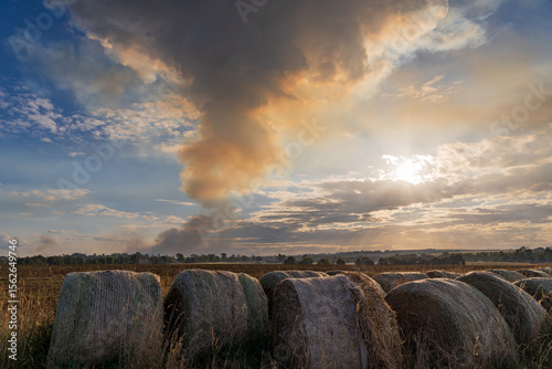 Smoke cloud rising at sunset over hay bales on farmland