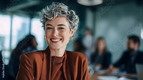 Smiling self assured woman with curly brown hair wearing a casual sweater standing in a bright contemporary office setting with coworkers in the background