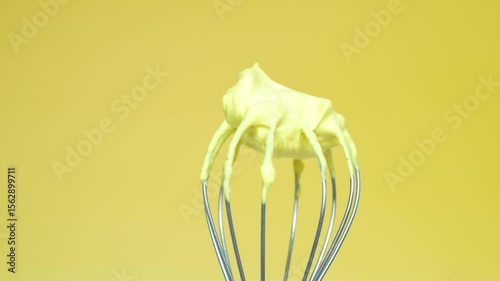 Whisk with yellow whipped cream from proteins close-up on yellow background.