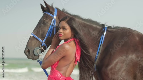 A young swarthy woman in a red dress stands next to a horse on the ocean shore.