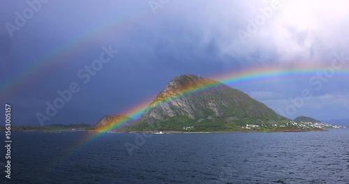 Bright rainbow arches over a lush mountain island in a Norwegian fjord, viewed from a cruise ship after rain. A stunning natural spectacle blending sea, sky, and landscape.