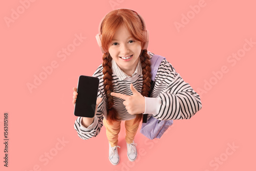 Cute ginger schoolgirl pointing at modern mobile phone with blank screen on p...