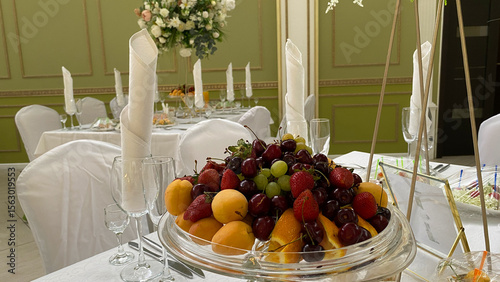 Beautifully arranged fruit platter featuring strawberries, cherries, and peaches on a glass dish. The elegant table setting with white linens and crystal glasses creates a perfect banquet atmosphere
