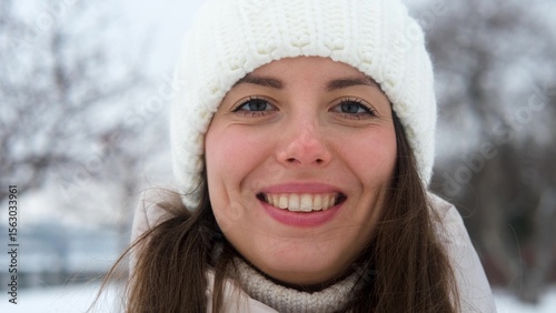 Portrait of woman in warm white knitted hat outdoors in winter. Young woman smiling and enjoying beautiful weather and first snow, laughing and looking at camera. Active winter recreation. Wide