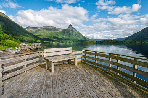 Tableau sur toile A wooden bench overlooks a calm fjord surrounded by lush greenery and majestic mountains under a partly cloudy sky