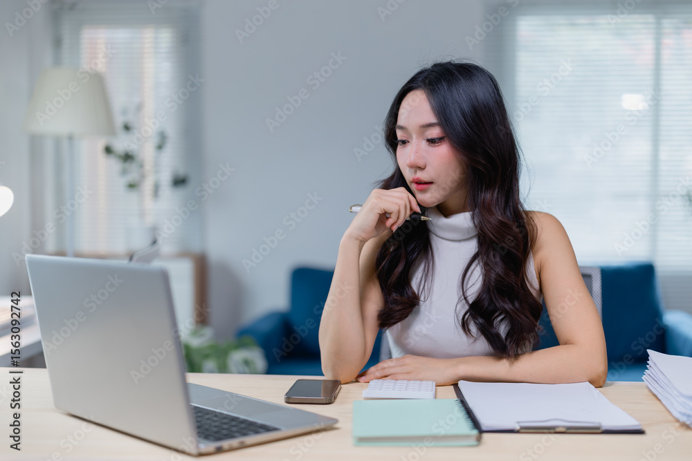 © Tj - Young woman working at a home office, focused on her laptop. She is surrounded by notebooks and a smartphone, indicating a productive and organized workspace