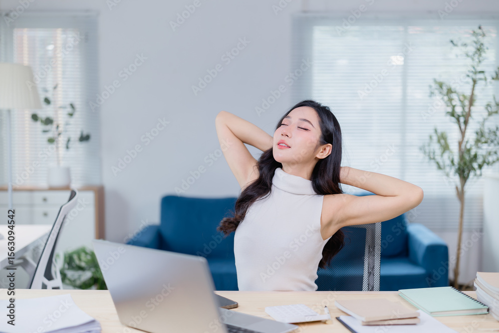 Obraz premium Woman takes a moment to stretch and relax at her desk in a modern office. She leans back with closed eyes, surrounded by a laptop, papers, and a comfortable workspace