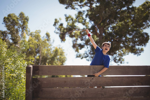 Boy leaning over wooden fence and raising red-wristbanded arm against clear sky in leafy park