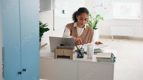 Female teacher flipping through pages in notebook and writing while sitting at desk and looking at laptop