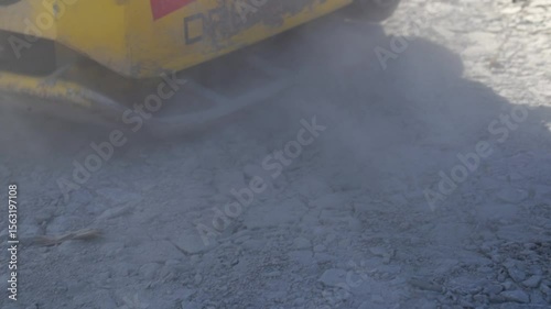 yellow construction machine compacting dusty gravel surface on road construction site with vibration and heavy equipment creating dust and flattening ground for building infrastructure and maintenance
