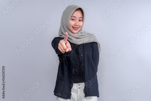 Smiling young Asian woman wearing a hijab, dressed in a black shirt, pointing forward with her index finger. The image conveys confidence, positivity, and an assertive attitude, ideal for branding.