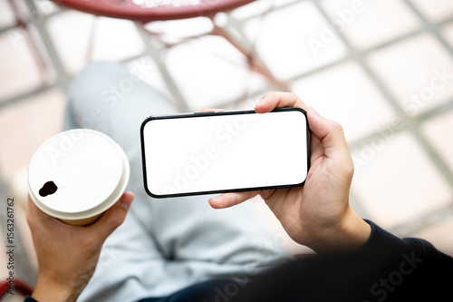 Hand holding coffee and white screen smartphone horizontally watching video playing game on bench.