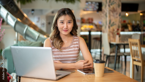 Pretty asian woman sitting with tablet and coffee beside laptop on wooden table in a cafe or canteen