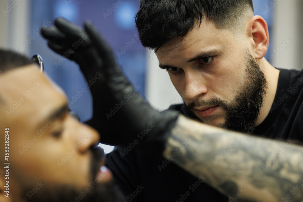 Obraz premium Barber shaving beard of client by using razor in barbershop