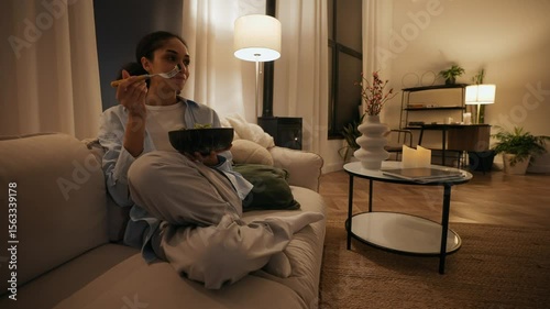 A woman sits on a couch and smiles while holding a fork to eat salad from a bowl