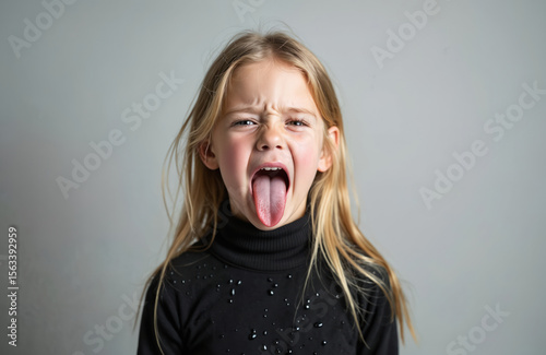 Young girl showing disgust face expression with tongue out. Blonde hair female child portrait in studio, grey background. Nonverbal communication of hate, dislike, disapproval. Negative feeling