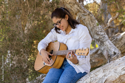 teeen girl sitting in shade of trees strumming a tune on an acoustic guitar