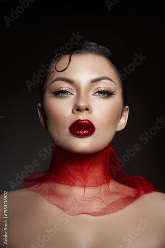 confident model poses, showcasing glossy red lips and dramatic eye makeup. sleek hairstyle complements the sheer red fabric around her neck, creating a bold and captivating look