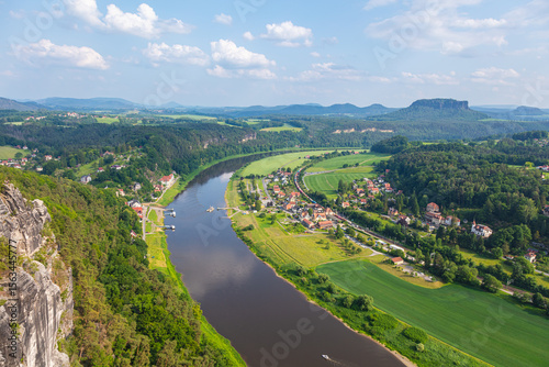 Scenic panoramic view from the Bastei cliffs over the Elbe River valley with sandstone rocks, lush forests, rural village rooftops, distant mountain ranges under a vibrant summer sky. Saxony, Germany