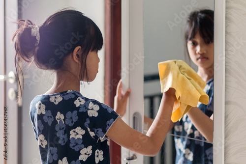 Household chores for children. A young Asian girl wiping the mirror in the wardrobe with a chamois cloth