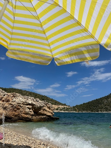 Yellow striped beach umbrella over a rocky shoreline with turquoise sea and hills in the background. Concept of summer vacation, seaside relaxation, and coastal tourism