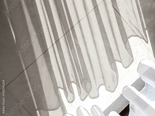 Sunlight streams through sheer curtains, casting long, dramatic shadows and playful wavy patterns on a textured light gray floor. A serene and minimalist abstract background.