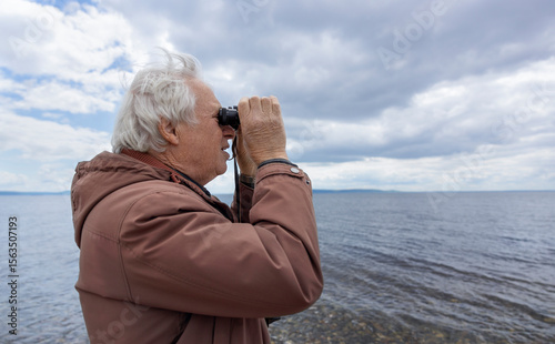 Senior man looking through binoculars at the beach under a thundery sky
