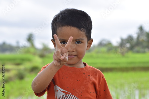 Southeast Asian child aged 3 smiling while holding up two fingers on blurred natural background