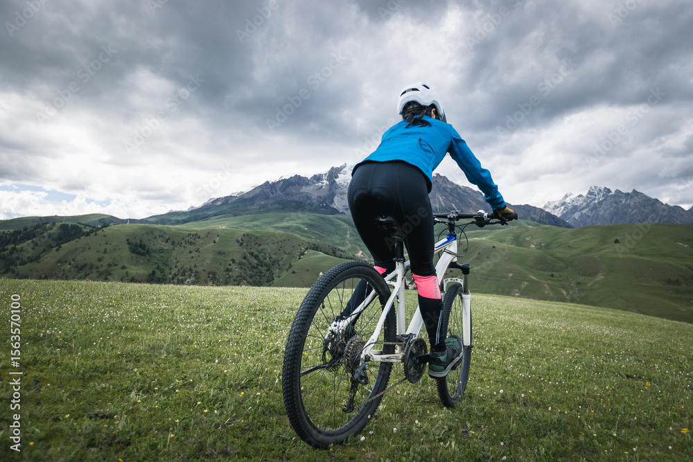 Obraz premium Woman cyclist riding mountain bike on grassland mountains with snow capped mountain in the distance