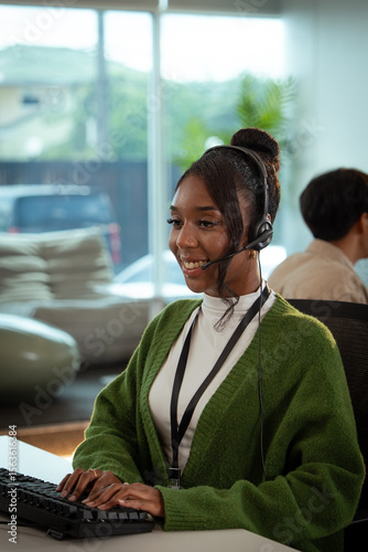 Cheerful customer support representative wearing headset and green sweater is working at desk in modern office environment, providing tech assistance