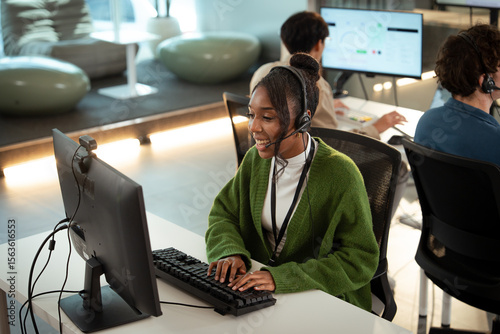 Fotografie Cheerful customer support representative wearing headset is working on computer in modern office environment
