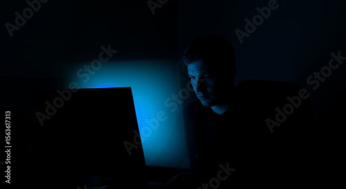 Late-night work session illuminated by a laptop's blue glow, a young man intensely focused on his task in the dark.