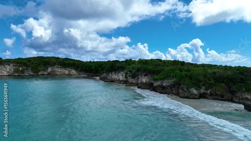 Incredible 4K drone footage showcasing a breathtaking tropical beach lined with palm tree-covered cliffs. Captured under a vibrant blue sky filled with soft white clouds