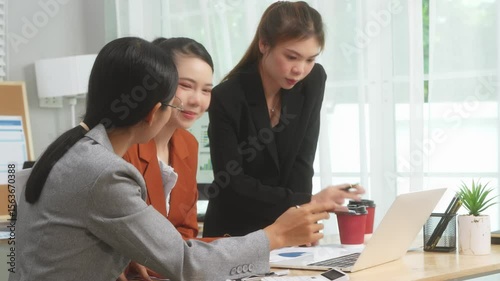 Three Asian women in formal suits collaborate at a desk, discussing documents in a modern office. teamwork, strategy planning, creative problem-solving in professional business environment.