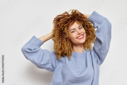 Smiling woman with curly hair wearing a light blue sweater, posing playfully against a neutral background, radiating joy and confidence in a modern lifestyle context