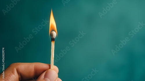 A close-up of a hand holding a burning matchstick against a soft teal background, capturing the vibrant flame and intricate details of the match.