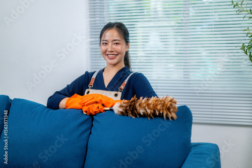 Housekeeper wearing orange gloves and apron smiling while cleaning a blue sofa with feather duster