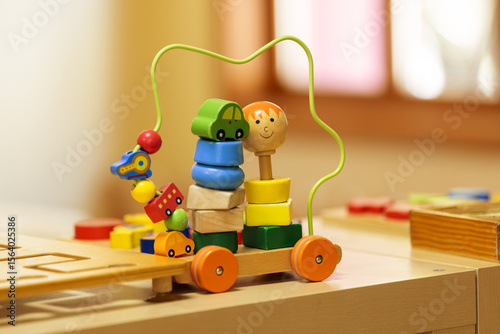 A colorful wooden bead maze and stacking toy with wheels sits on a light wooden surface in a kindergarten setting. The image highlights engaging educational play for young children.