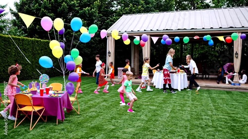 Excited kids racing through garden during birthday celebration, colorful balloons surrounding playful moment with adults preparing festive meal