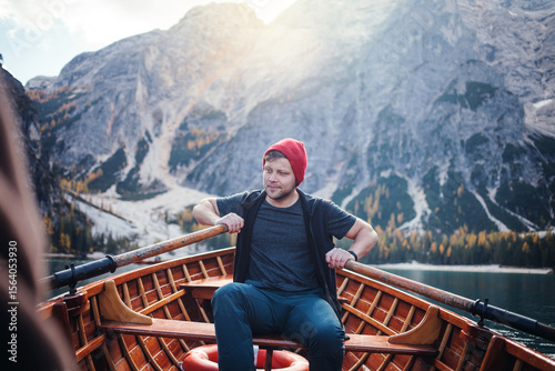 A man in a red beanie sits in a wooden rowboat on a clear lake, surrounded by majestic mountains and a sunny sky