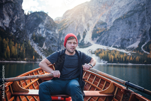 A man in a red beanie sits in a wooden rowboat on a clear lake, surrounded by majestic mountains and a sunny sky