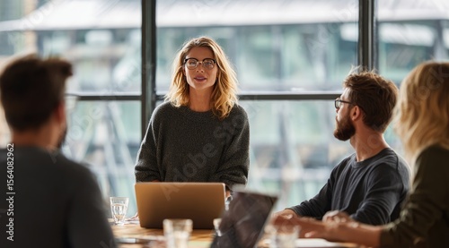 A woman leads a small group meeting around a table, presenting to attentive colleagues near a large window