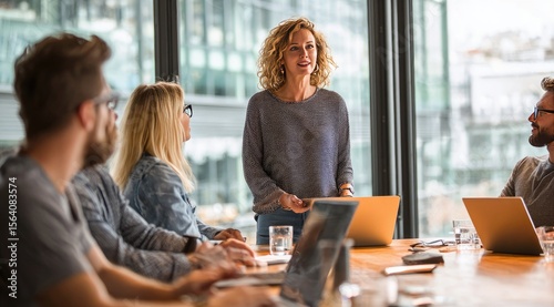 A woman leads a team meeting, presenting information to colleagues seated around a large table with laptops and glasses of water.  Natural light streams in from a large window behind her